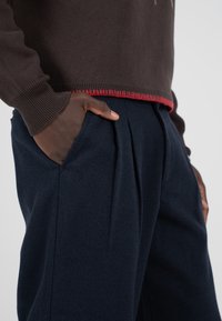 Brązowy sweter z dzianiny z czerwonym wykończeniem u dołu, w parze z granatowymi spodniami plisowanymi z bocznymi kieszeniami. Miękka tekstura i luźny krój.