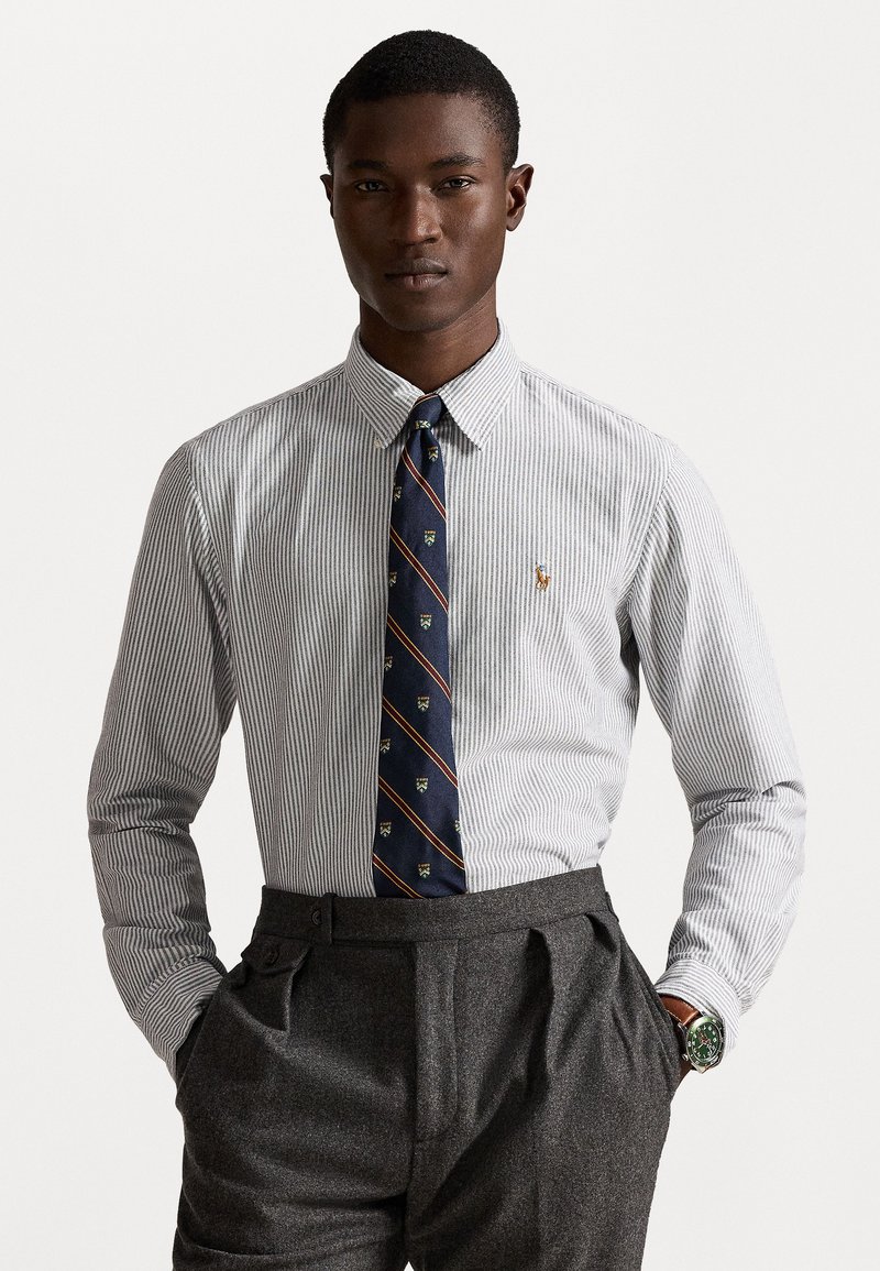 Balta un tumši zila svītrainā krekls ar pogājamām apkaklēm, izšūts logo, komplektā ar tumši zilu kaklasaiti ar krāsainām apdrukām, pelēki noderīgi uzvalka bikses.