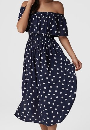 Femme portant une robe bleu marine à épaules dénudées avec des pois blancs, taille smockée et jupe fluide, tenant un côté de la robe.