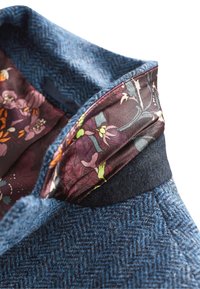 Manteau en laine présentant un motif en chevrons bleu, avec une doublure bordeaux à motif floral visible au niveau du col. Texture douce et design ajusté.