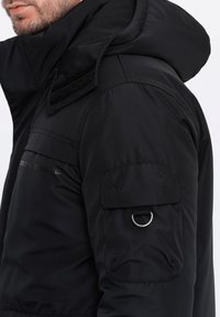 Černá vycpaná bunda s kapucí, se strukturovaným předním dílem, suchými zipy na límci a kapsou na nářadí s kovovým kroužkem.