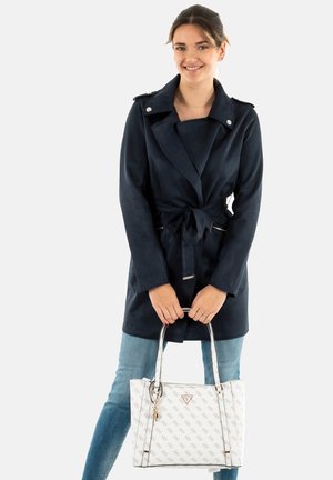 Manteau trench bleu marine avec taille cintrée, col en revers et accents argentés. Sac à main blanc avec motif de logo répété. Jean en denim.