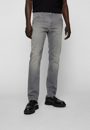 Homme portant un jean gris slim et des mocassins en cuir noir, debout devant un fond clair uni.