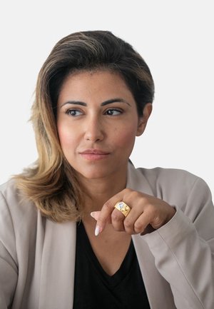 Anello d'oro con una grande pietra trasparente, forma esagonale, indossato sulla mano destra. La persona ha capelli scuri di lunghezza media e indossa una giacca beige chiaro. Sfondo bianco.