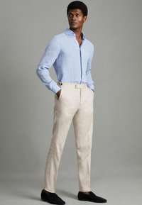 Chemise en lin bleu clair à manches longues avec col, associée à un pantalon crème à front plat et détails de boutons, portée avec des chaussures noires.