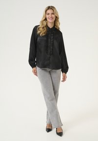 Blusa de satén negra con detalle de volante en el frente, mangas largas y cierre de botones. Combinada con jeans grises y tacones negros de punta.