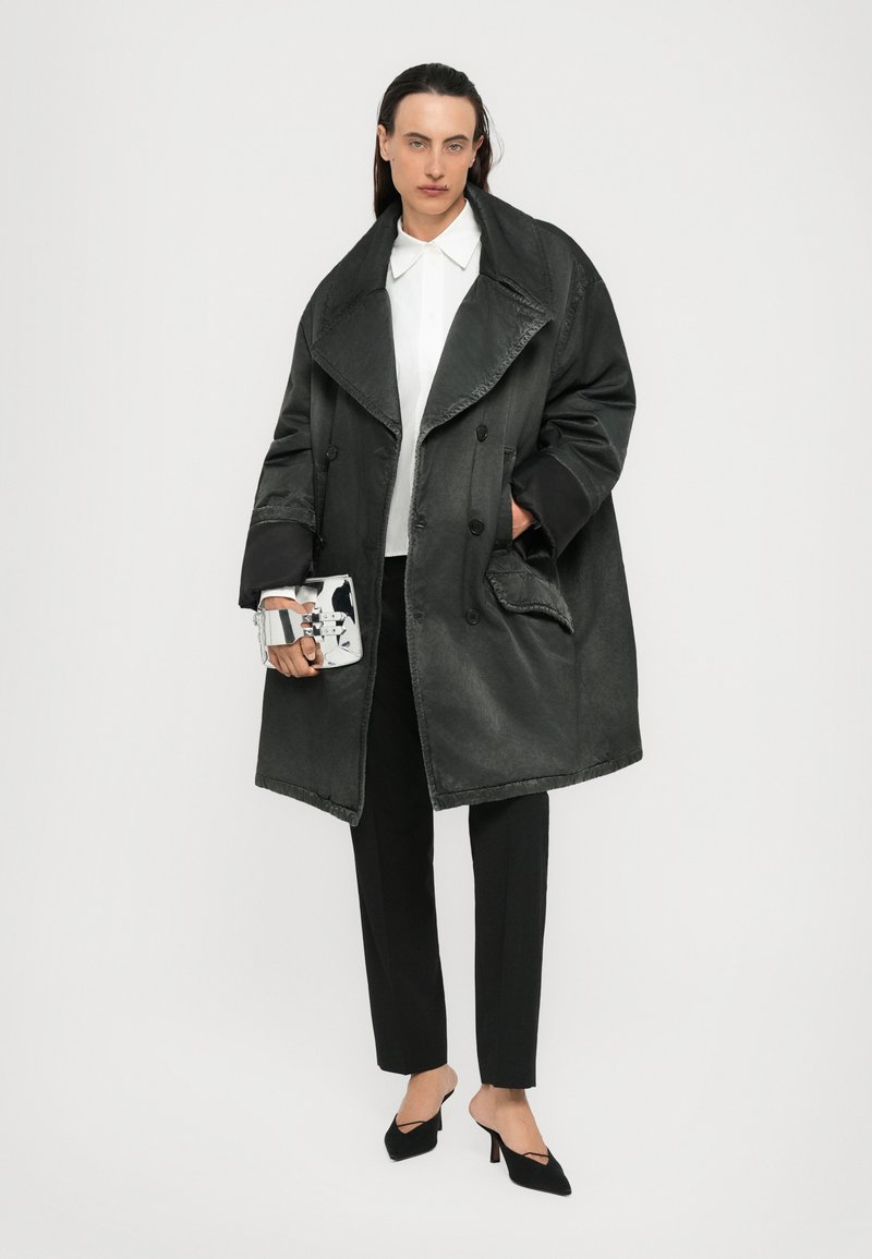 Personne portant un manteau double boutonnage gris foncé surdimensionné, une chemise blanche à boutons, un pantalon noir, des talons noirs, tenant une pochette réfléchissante argentée.