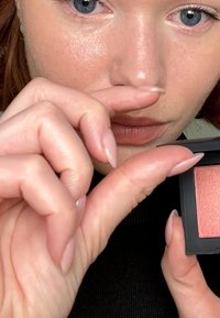 Blush compacto en un pequeño estuche negro; presenta un suave tono coral brillante. La modelo muestra el ancho de su dedo para indicar su tamaño.