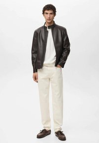 Blouson en cuir marron avec un col haut, une fermeture éclair et des poignets élastiques, associé à un pantalon crème et des chaussures à lacets marron.