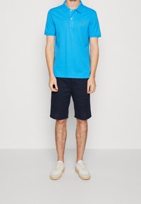 Camisa polo azul de manga corta, con cuello y un pequeño logo. Combinada con pantalones cortos de un oscuro color marino y zapatillas blancas.