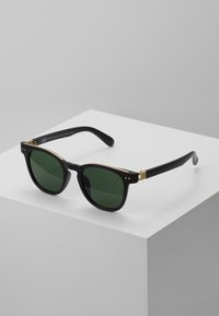 Black sunglasses with green lenses, featuring round lenses, thick frames, gold accents near the hinges, and a matte finish on the arms.