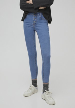 Person iført højtsiddende blå skinny jeans med fire knapper, mørkegrå sokker, hvide sneakers og en sort langærmet top med armene krydset.