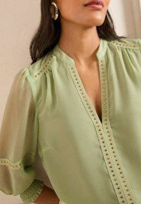 Blusa verde chiaro con maniche a sbuffo trasparenti e corte, caratterizzata da borchie in tono oro lungo la patta e le cuciture delle spalle. Tessuto dalla texture liscia.