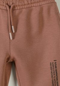 Pantalon en maille marron avec taille élastique et cordon de serrage, comportant un texte noir sur la cuisse droite affichant un message motivant.