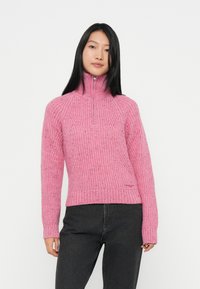 Rosa strikket genser med høy krage, med ribbestrikkede mansjetter og halskant, samt et strukturert mønster. Kombinert med mørkegrå bukser.