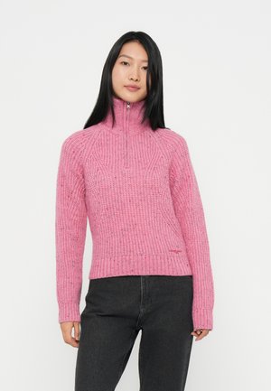 Rosa gestrickter Pullover mit hohem Kragen, mit gerippten Bündchen und Saum sowie einem strukturierten Muster. Kombiniert mit dunkelgrauen Hosen.