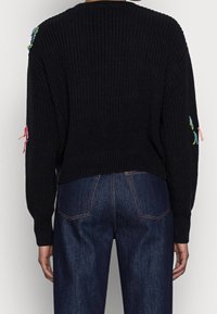 Černý pletený svetr s volným střihem a žebrovanou strukturou, zdobený barevným vyšíváním a střapci na ramenou, kombinovaný s tmavými džínami z denimu.