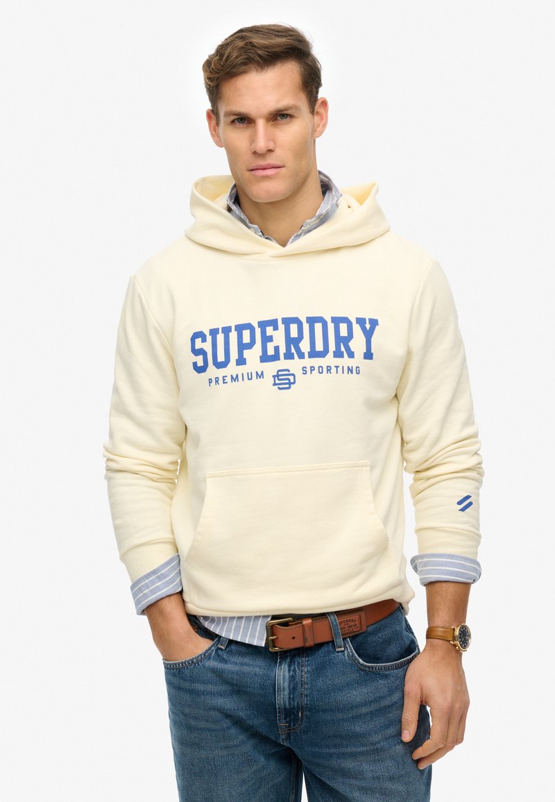 Sweat à capuche crème avec logo "SUPERDRY" bleu. Comprend une poche avant, des poignets côtelés, et une chemise superposée à rayures bleues en dessous.