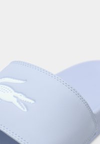 Sandale à slide bleu clair avec une bride texturée affichant un logo crocodile en relief. Dessus en matériau lisse avec des détails couture.