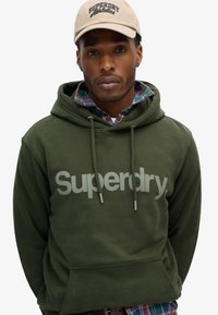 Oliven grøn hættetrøje med et stort gråt "Superdry" logo, snøre til hætten og forlommekænguru, lagdelt over en ternet skjorte.