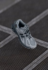 Harmaa juoksukenkaat, joissa on verkkomateriaalista päällinen, mustat yksityiskohdat, pehmustettu kaulus ja teksturoitu pohja, jossa on korostettu pito. Varustettu nauhoitusjärjestelmällä tukevan istuvuuden saavuttamiseksi.