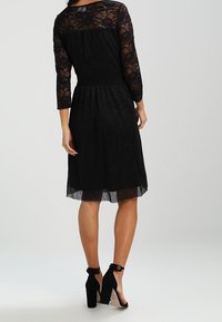 Musta pitsimekko, jossa on kolmanneksen mittaiset hihat, teksturoidut hameet ja polvipituinen helma. Yhdistetty mustiin korkeakorkoisiin kenkiin, joissa on nilkkahihnat.