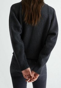 Personne aux longs cheveux portant un pull en maille gris foncé et un jean sombre, debout les mains jointes dans le dos sur un fond uni.