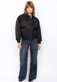 Must must jacket, millel ukse ees, küljetaskudega ja soonikkoes varrukatega. Materjal on sile ja läikiv, rinnal on logotäht.