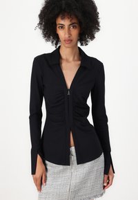 Blusa nera a maniche lunghe con colletto, zip frontale, dettagli arricciati e polsini con spacco laterale, realizzata in tessuto morbido.