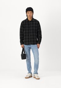 Chemise à carreaux noir et gris, boutonnée, assortie avec un jean bleu clair. La personne tient un petit sac en bandoulière noir et porte des baskets blanches.