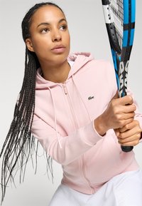 Haut de survêtement zippé rose clair avec cordons de serrage, arborant un petit logo vert. Le mannequin tient une raquette de tennis bleue et noire, portant un short de sport blanc.