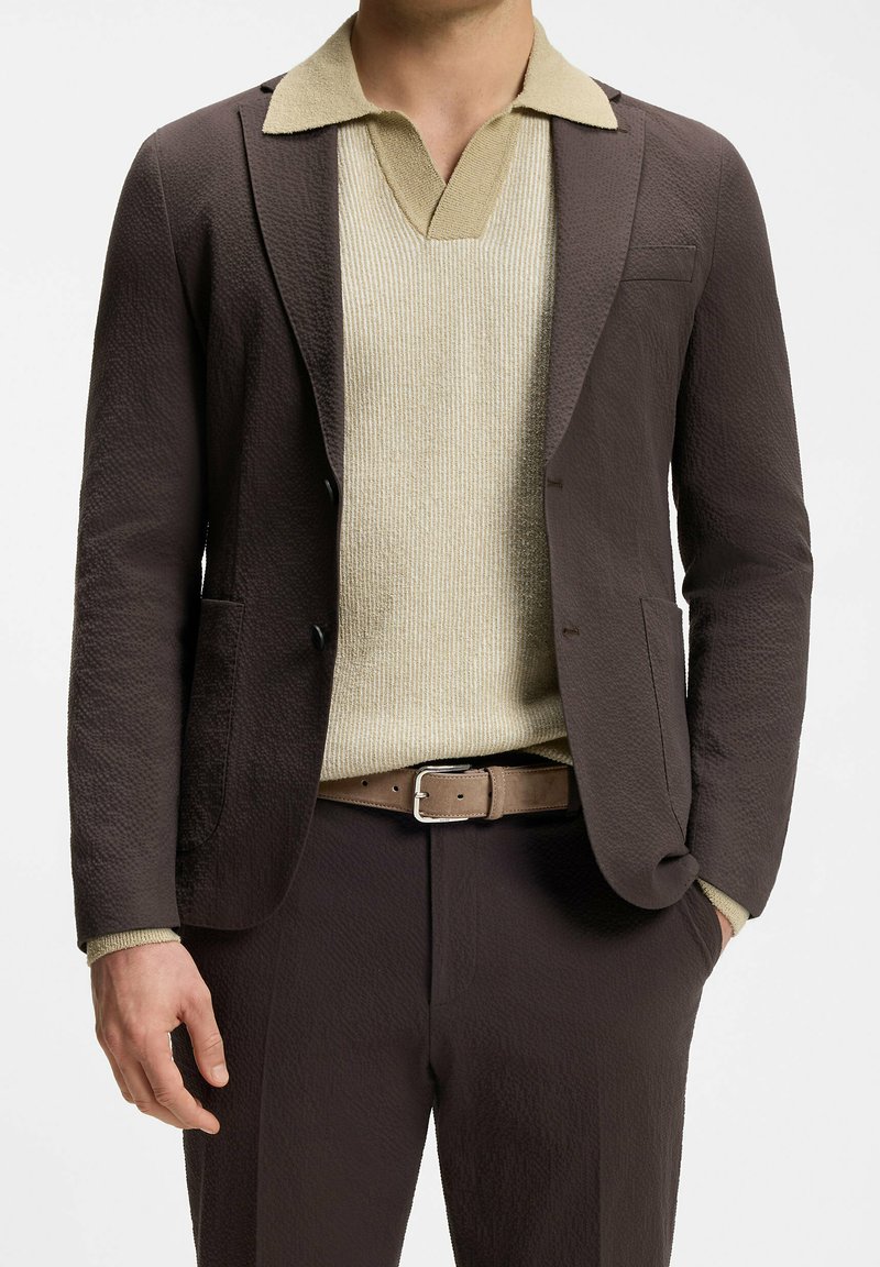 Uomo con un completo marrone scuro dalla texture a rilievo, indossa una polo lavorata a maglia beige infilata nei pantaloni, con una cintura beige, una mano in tasca.