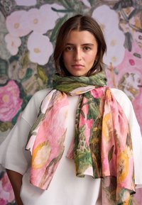 Květinový šátek zdobený růžovými a žlutými květy s zelenými akcenty, vyrobený z lehké látky, přehodil se kolem krku, na pozadí s květinovým vzorem.