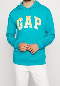 Sudadera con capucha turquesa con el logo "GAP" en amarillo, hecha de mezcla de algodón, cuenta con un bolsillo tipo canguro en el frente y capucha con cordón. Textura suave.