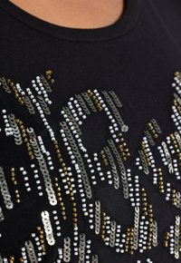 Primer plano de una tela negra con lentejuelas decorativas en plata, oro y blanco, formando un patrón ondulado en la superficie.