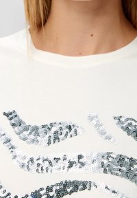 Vit T-shirt med ett mönster av silver- och grå paljetter arrangerade i vågor, med en mjuk textur och rund halsringning.