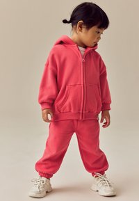 Pink hættejakke med lynlås og matchende joggingbukser, begge i blødt stof. Har elastiske manchetter og lommer, kombineret med hvide sneakers.