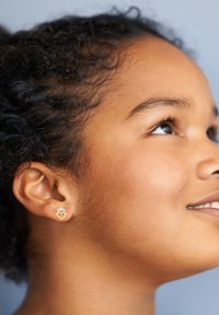 Guldörhängen i hjärtform med en krondetalj, med en slät finish, bärs av en person med lockigt hår mot en enfärgad bakgrund.