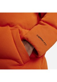 Oranger Puffer-Jacke mit einer Stoffstruktur. Verfügt über eine Seitentasche und ein schwarzes, gesticktes "THERMA-FIT"-Logo am Ärmel.