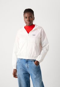 White zip-up windbreaker with hood, featuring a front pocket and "Tommy Jeans" logo. Paired with light blue denim jeans.