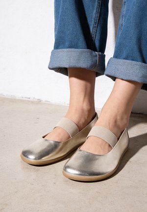 Piedi con ballerine Mary Jane argento metallizzato con cinturini elastici, abbinate a jeans di denim blu risvoltati su un pavimento chiaro.