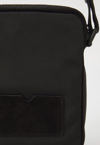 Must mustriga üle õla kott, millel on esiküljel tekstuuriga suedepatch, lukuga kinnitamine ja reguleeritav rihm.