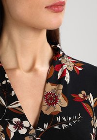 Ženská dolní část obličeje a krku s černým oděvem s květinovým vzorem, na kterém jsou červené, béžové a bílé květiny a listy.