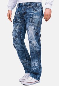 Jeans di denim con un lavaggio blu sbiadito, caratterizzati da più tasche, motivi cuciti e una vestibilità ampia. Completati da sneaker bianche.