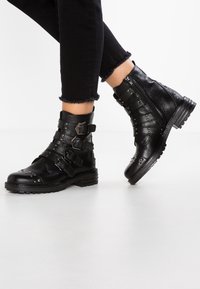 Černé kožené kotníkové boty s několika pásky, stříbrnými hroty a bočním zipem. Texturovaný povrch s robustním podpatkem a gumovou podrážkou.