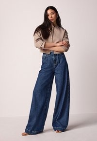 Giovane donna con lunghi capelli scuri che indossa una giacca corta beige e jeans a gamba larga blu scuro, in piedi con le braccia incrociate.
