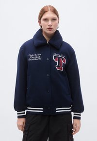 Tamsiai mėlyna varsity stiliaus striukė su pūsta apykakle, siuvinėjimais ir 'T' lipduku, papuošta baltomis dryžuotomis rankogalių ir juosmens juostelėmis.