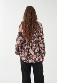 Blusa nera fluida con motivo paisley floreale in rosa e viola, maniche lunghe, vestibilità rilassata e orlo curvo. Indossata con pantaloni neri.