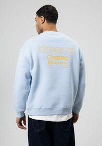 Sweat-shirt bleu clair à la coupe décontractée ; un texte jaune vif au dos indique « dreams Chasing NOT deadlines ». Manches et bas côtelés.