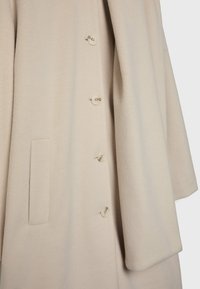 Manteau en laine beige présentant une texture douce, de gros boutons et une poche latérale ; conçu avec une coupe décontractée et des manches larges.
